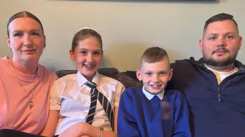 Mum, Kylie, wearing a pink T-shirt, their 13-year-old daughter Casey, wearing her white school shirt and blue striped tie, nine-year-old son James, wearing a blue school jumper and dad Lee, wearing a blue zip-up jumper - sitting together on a sofa smiling at the camera.