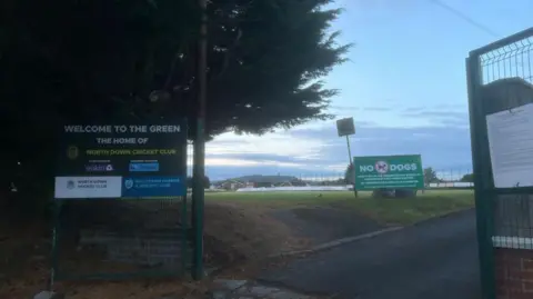 A view of North Down Cricket Club through the open gates. A sign says 'Welcome to the Green, the home of North Down Cricket Club.' A sign further back says: 'No dogs'
