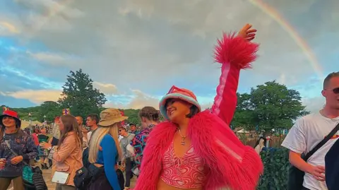 Lucy Smith-Butler At another festival, Lucy is smiling with her eyes closed and arm in the air amongst a crowd of people. Her outfit is bright pink, including a jacket with a furry trim and a pink bucket hat. A rainbow is visible in the sky above her.