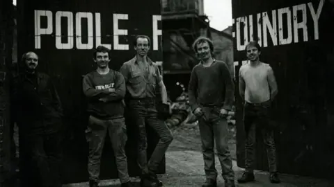 Gary Norman Black and white photo of five men posing in front of the open gates. The words 'Poole Foundry' are written above their heads across the gates.