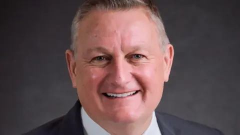 NAHT Head teacher Simon Kidwell smiles into the camera.  He has gray hair and is cleanshaven.  He wears a white shirt and a dark gray jacket