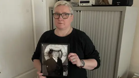 BBC A woman with short blond hair and glasses holds a black and white picture of her younger self in a Navy uniform