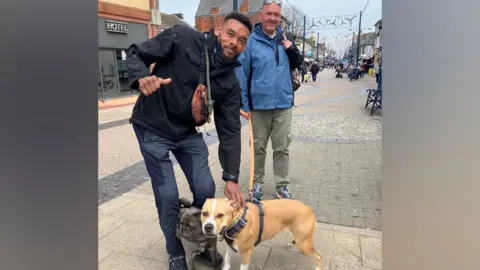 Owen Sennitt/BBC A man crouches down to stroke two dogs and puts his thumb up, while another man in a blue jacket smiles towards the camera