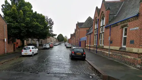 A general view of the entrance to Edward Street in Carlisle. The cobbled street has red-brick buildings either side of the road, with cars parked outside the properties.
