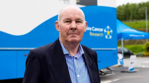 A man in a black jacket and blue and white stripe shirt stands in front of a mobile scanning unit. 