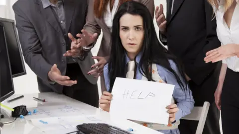 Getty Images Woman sitting among business colleagues holding up paper with word 'help' written on it