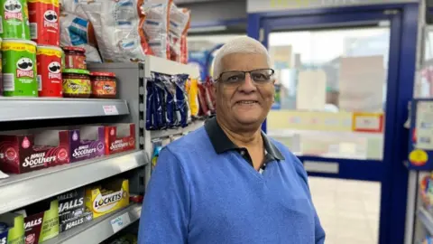 Ollie Conopo/BBC Suresh Patel with short white hair and glasses in a small local shop