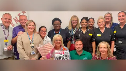 North West Anglia Foundation Trust  Mary Donaldson surrounded by her colleagues who are all smiling at the camera. Mary is pictured with a bouquet of sunflowers, leaving gifts and a large leaving card which is designed to look like a tabloid paper. She has blonde hair and is smiling at the camera.