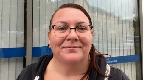 A woman with brown hair in a ponytail standing in Bargoed high street. She is wearing glasses and smiling at the camera. It is a head and shoulders shot and she has a window with white blinds behind her.