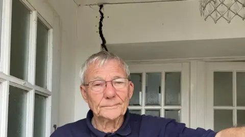 Alexander Stevanovic/BBC Roy is an older gentleman, with grey hair, wearing glasses and a navy polo shirt. He is standing on the porch of his home with a big crack in the wall behind him, which is caused by the subsidence.
