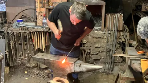 Simon Hall is at work looking down at a poker which has a tip glowing white and orange with heat resting on an anvil. His right hand is bringing a hammer down onto it. Behind him is a furnace and the tools of his trade.