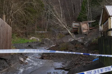 Police tap cordons off a street, there are mounds of rubble and mud. There is debris from small sheds as well as water coming down the road