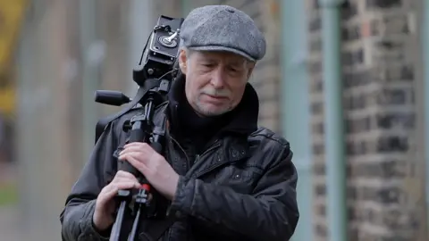 Jason Thompson Mik Critchlow is carrying a camera over his shoulder. He has grey stubble and is wearing a black coat and has a grey cap on. He is walking alongside a row of cottages 