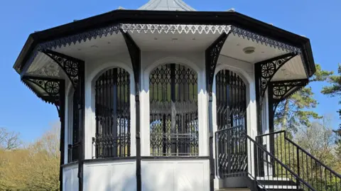 A close-up of the black and white bandstand in Bedford Park that has been refurbished. It is set against a bright, blue sky.