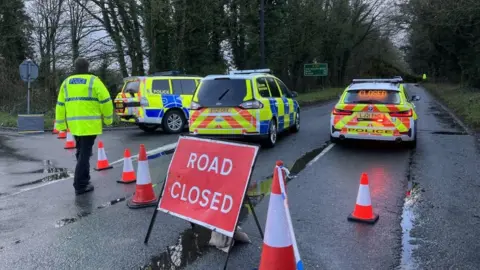 Three police cars at a scene with roads closed signs