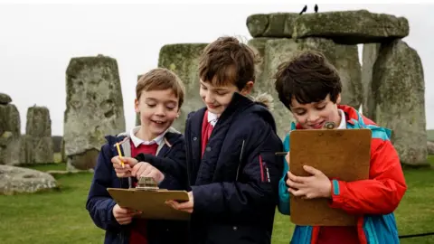 English Heritage A group of three schoolchildren with clipboards in front of Stonehenge