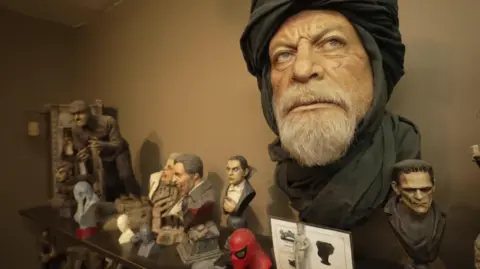 Hill's mask designs on his shelves in his LA studio including a large head design featuring actor Oliver Read alongside smaller models of Frankenstein, Dracula and Spiderman.