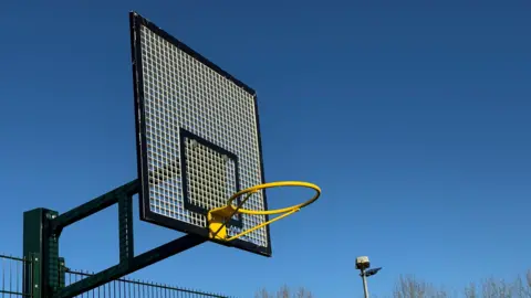 North Northamptonshire Council A basketball hoop, with a blue sky around it, the hoop is yellow and there is a flood light to the right. 