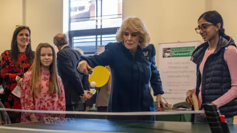 Queen Camilla playing table tennis with a yellow paddle. She is wearing a blue coat and a broach. Next to her are two children watching as well as a woman in a red dress.