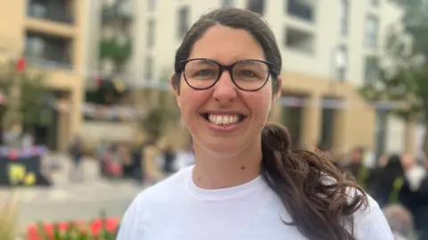 Vivianne is wearing glasses and a white “LUSO Project Luso” T-shirt standing in a modern outdoor plaza, with blurred apartment buildings, bunting, and people in the background.
