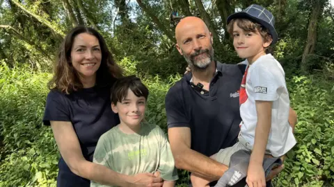 A family of four pose in woodland, with one child standing in front of an adult and another being held. A treehouse is visible among the trees in the background.