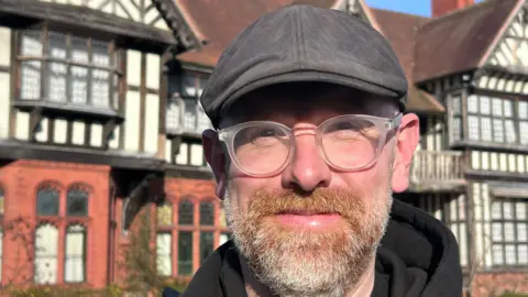 James Carnell A man with a grey beard and white glasses and a black flat cap is looking at the camera with a slight smile and historic house in the background