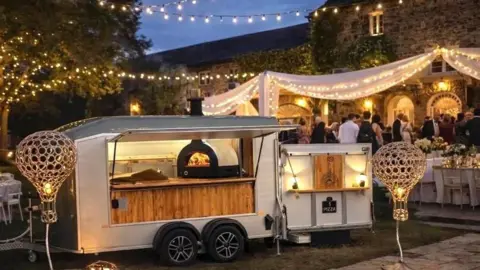 Copper Kiln Pizza A mobile wood fired pizza van which is white and has a hatch that is open. It is at an event that looks like a wedding, with a dark sky and fairy lights strung up between trees. There is a tent with fabric draped across the top