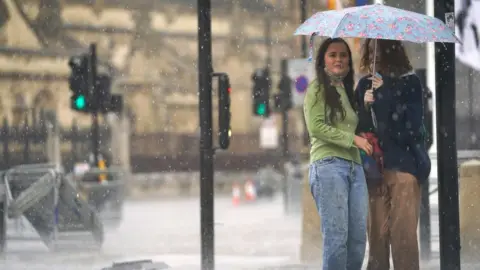 PA Media Two young women shelter under an umbrella in Parliament Square in central London amid heavy rain