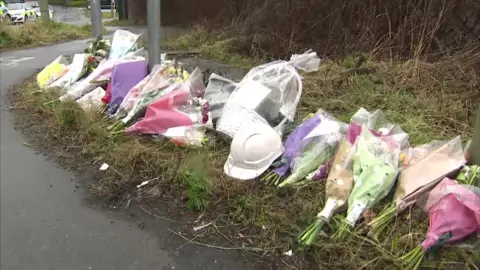 A number of bouquets of flowers, gifts and helmets that buy together to the victims of the plants. It is a cloudy day. Police cars can be seen in the distance.