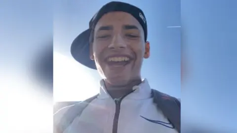 Cameron Bailey giving a big smile/laugh against a blue sky. He is wearing a baseball cap with the peak on his right side and a white Nike hoodie, with rucksack straps over his shoulders.