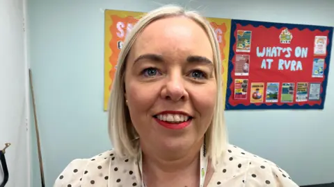 A woman with shoulder length blonde hair, wearing a white top with black dots on it. There is a red poster on the blue wall behind her, reading 'what's on at RVRA'. 