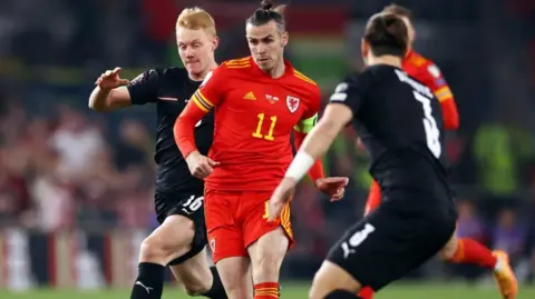 Gareth bale in Wales kit dribbling between two austrian players in black kit. Gareth has an intent look and has his usual Bale bun ton knot hairstyle 