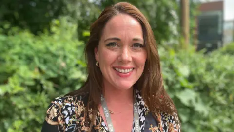 A white woman with brown hair standing in front of a hedge smiles toward the camera. She is wearing a black shirt and a grey lanyard with staff written on it.
