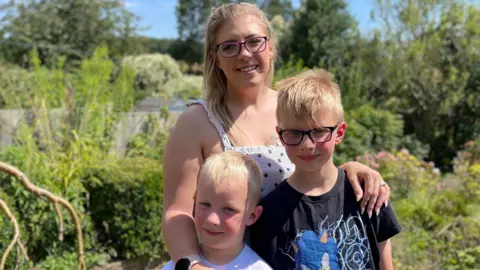 Alicia with her two sons. She has long blonde hair and is wearing a white dress with black spots in a garden with plants behind her. Her sons both have blonde hair, one is a foot taller than the other and wears black glasses and a black T-shirt with a Sonic the Hedgehog logo while the other has a white T-shirt