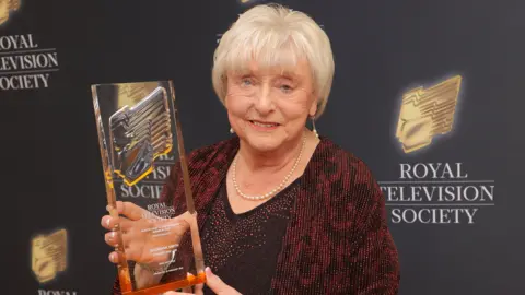 Ann Ming holds a large glass trophy in front of a black banner with the Royal Television Society written on it. She is elegantly dressed in a dark red and black jacket, with short white hair.