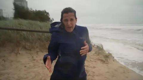 Katy Watson on a beach wearing a rain coat with hair blown in strong winds