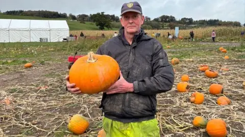 A man dressed in a muddy black coat and hi-viz overalls standing in a field sporadically covered with pumpkins while holding one in his arms.