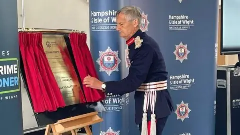 Hampshire & Isle of Wight Fire and Rescue Service Lord Lieutenant Nigel Atkinson unveils a plaque from behind red curtains at the new station. He wears his formal blue uniform.