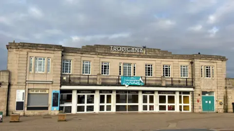 Old seaside building on the seafront. It is a classic opening to an old lido on a road. There is a big sign at the top that says "Tropicana" with a cloudy, moody sky.