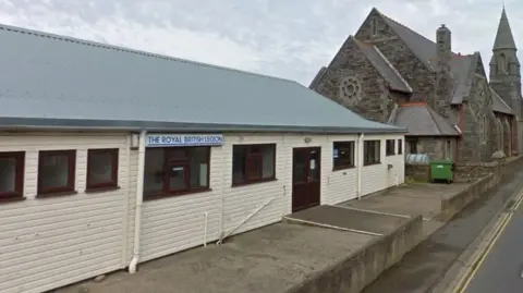 The Port Erin Royal British Legion building, which is a cream coloured, panelled, single storey building with brown framed windows and doors and a grey sloping roof. There is a stone build church building to the right of it.