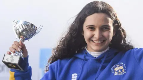 Stephanie wearing a blue hoodie with long curly brown hair smiling at the camera. She is holding an award.