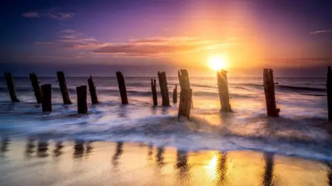 Chris McLoughlin / Getty Images A purple and orange sunrise at Spurn Point in East Yorkshire. Waves crash against the old coastal defences.