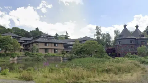 Powys County Council's headquarters in Llandrindod Wells. It is a two-storey brown brick building with a dark-coloured tiled roof. There are several buildings of the same style surrounding it and they sit next to a pond with trees dotted around.