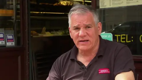 Chris Georgiou, an older man in a black uniform, speaking outside his cafe Speedy's.