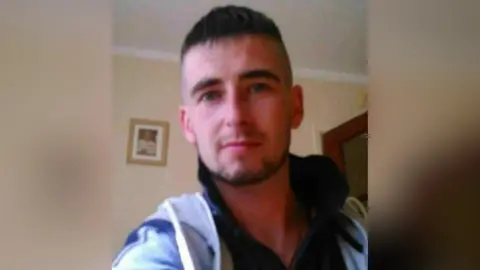 Family handout A picture of Dominic McGrath. He is a young man with short, dark hair and a beard. He is wearing a dark polo shirt and a light blue zipped hoodie. He is standing in a pale coloured room and is smiling at the camera.