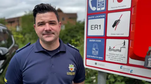 A blue uniformed fire service officer stands beside a waterside safety station, which includes instructions for using an emergency throw line. The equipment panel features symbols showing how to call 999 and rescue someone from the water. The man has short brown hair, and a Humberisde Fire & Rescue insignia on his shirt.