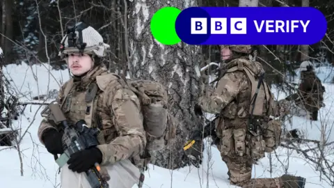 UK troops on Nato cold weather exercises in Estonia. Two soldiers can be seen in the snow, wearing camouflage gear and helmets and holding rifles. 
