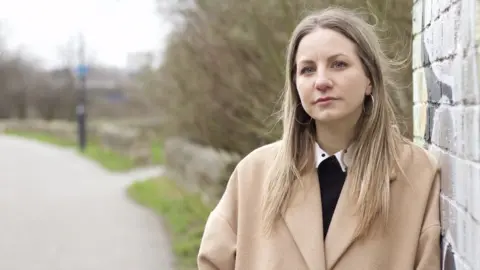 Sarah stands outside by a public footpath, by a wall