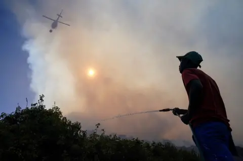 NIC BOTHMA/REUTERS A man and a helicopter pour water to put out a fire.