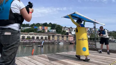 A man is wearing a yellow and blue costume made to look like a zero percent beer can with the name Clear Head. He is holding a hand glider above his head at the edge of a raised platform looking down onto Bristol Harbour.